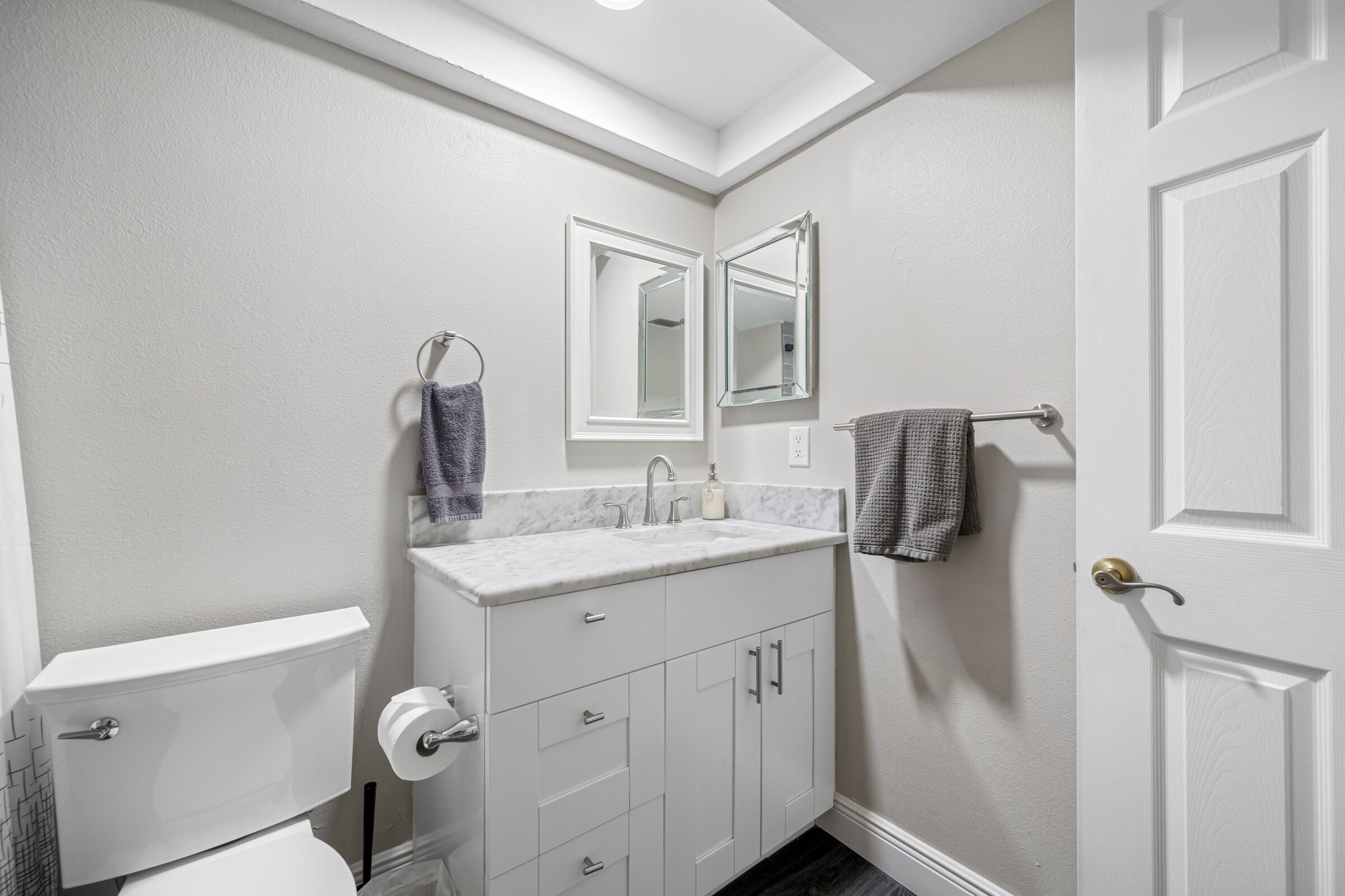 A bathroom with a white toilet, a sink with a marble countertop, and a mirror above it. There are towels hanging on a rack and a door with a brass handle.