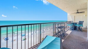 A balcony with a view of the beach and ocean, a ceiling fan, and outdoor furniture.