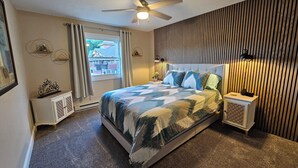 A bedroom with a bed, a ceiling fan, a window with curtains, a bedside table with a lamp, and a wall with vertical wooden slats. - Cambrian & Co. Downtown Loft Stylish, Sophisticated, Charm (Taylors Falls)