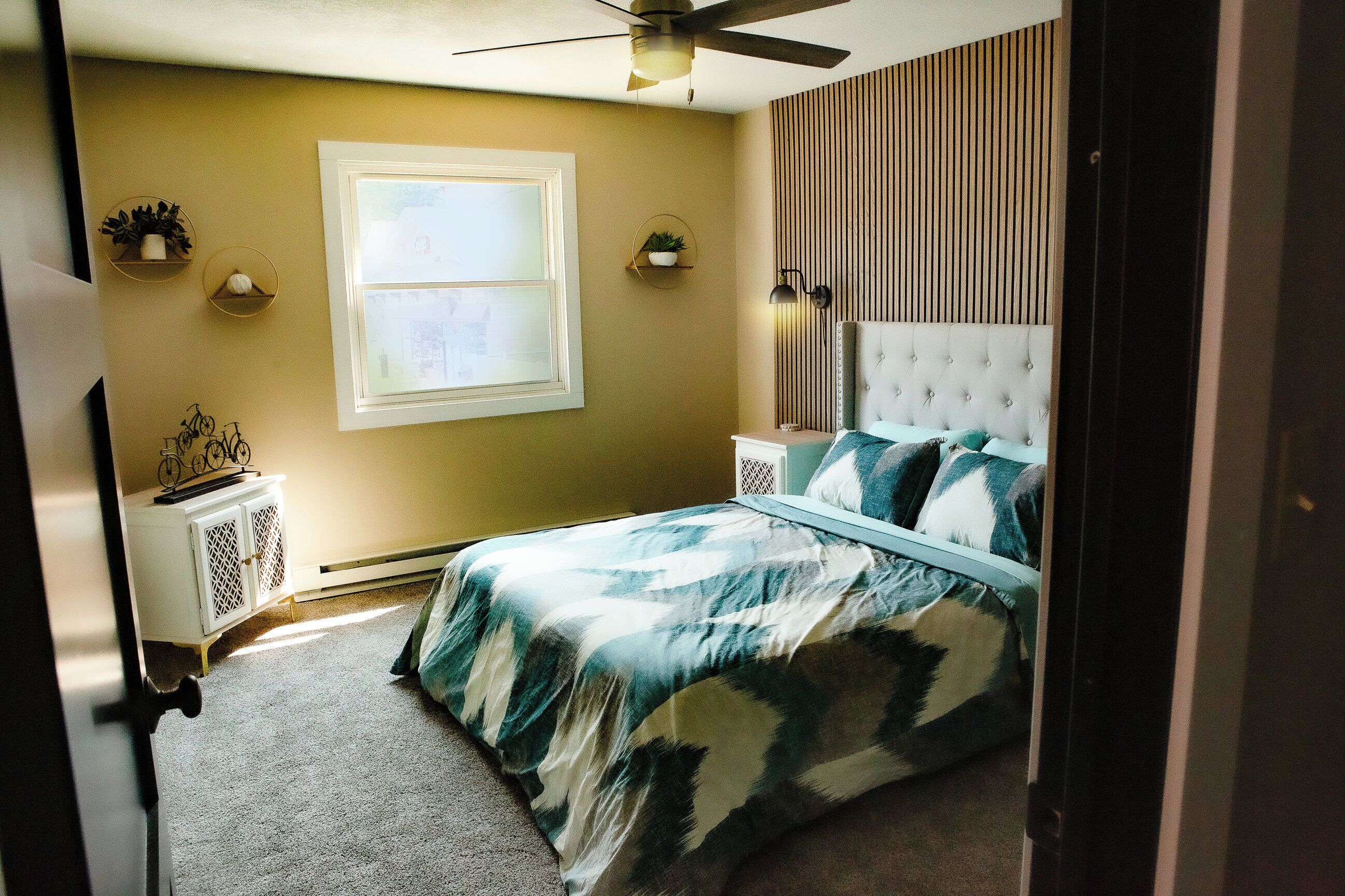 A bedroom with a bed, a window, a ceiling fan, and a nightstand with a lamp.