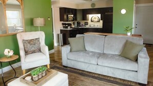 A living room with a grey sofa, a white armchair, and a small round table with a lamp. A kitchen is visible in the background. - Cambrian & Co. Downtown Loft Stylish, Sophisticated, Charm (Taylors Falls)