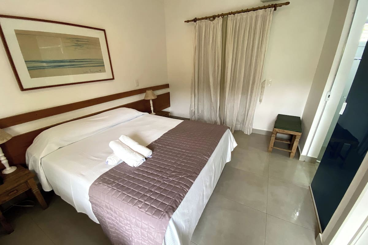A neatly made bed with a brown quilt, white linens, and rolled towels. There is a wooden nightstand with a lamp, a small wooden stool, and a framed picture on the wall.