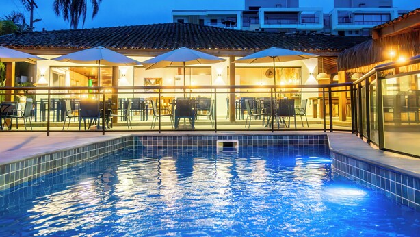 A swimming pool with lounge chairs and umbrellas, surrounded by buildings with balconies and a tiled roof. - MAR E PRAIA HOTEL (Ubatuba)