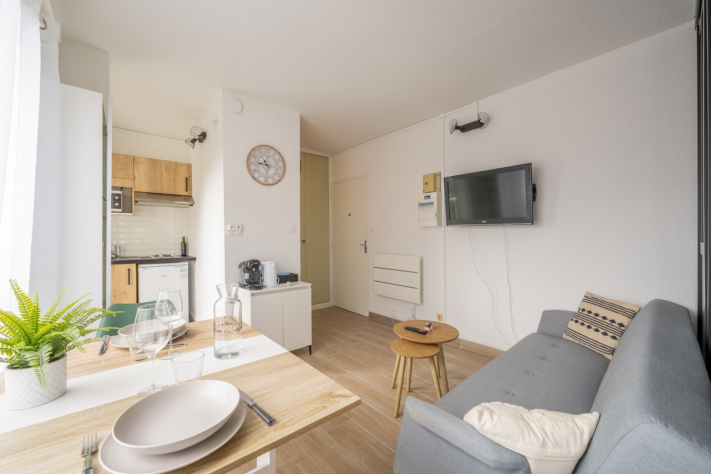 Ein modernes Wohnzimmer mit einem grauen Sofa, einem kleinen Holzesstisch mit Tellern und Gläsern, einer Küche im Hintergrund und einem an der Wand befestigten Fernseher.