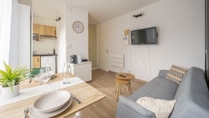 Ein modernes Wohnzimmer mit einem grauen Sofa, einem kleinen Holzesstisch mit Tellern und Gläsern, einer Küche im Hintergrund und einem an der Wand befestigten Fernseher.