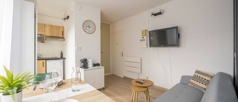 Ein modernes Wohnzimmer mit einem grauen Sofa, einem kleinen Holzesstisch mit Tellern und Gläsern, einer Küche im Hintergrund und einem an der Wand befestigten Fernseher.