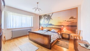 A bedroom with a large bed, a nightstand, a chair, a table, a window with curtains, a radiator, and a wall painting of a sunset over palm trees.