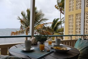 Una mesa dispuesta con un vaso de jugo de naranja, un cuenco de fruta y una taza, con vista a una masa de agua con palmeras.