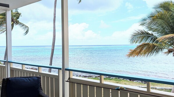 A balcony with a view of the ocean, palm trees, and a chair.