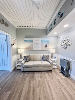 A living room with a beige sofa, two side tables, a fireplace, and a wall with decorative items. - Beach Haven, Only 5-Star Reviews, 1st Floor Condo! (Myrtle Beach)
