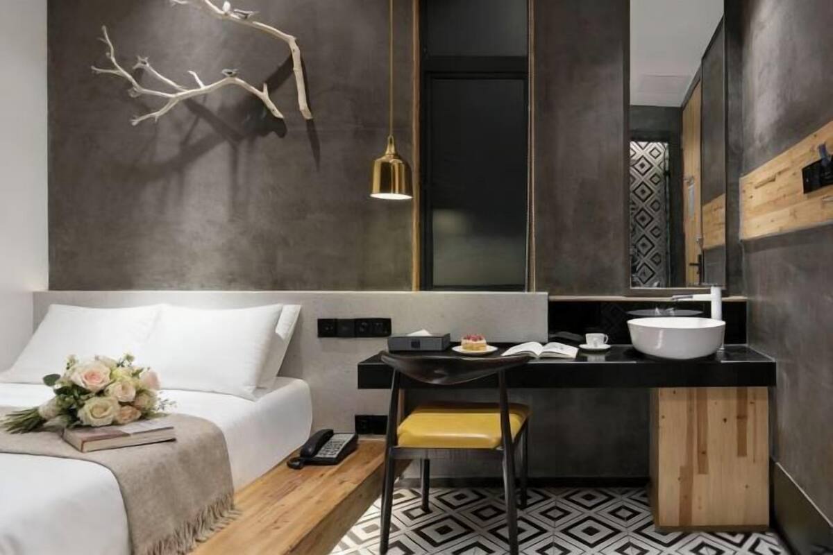 A modern hotel room with a bed, bedside table, chair, and a small desk with a bowl and books.