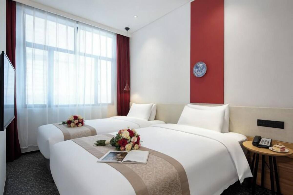 A hotel room with two beds, a red accent wall, a clock, a bedside table with a phone and a plate of food, and a window with curtains.