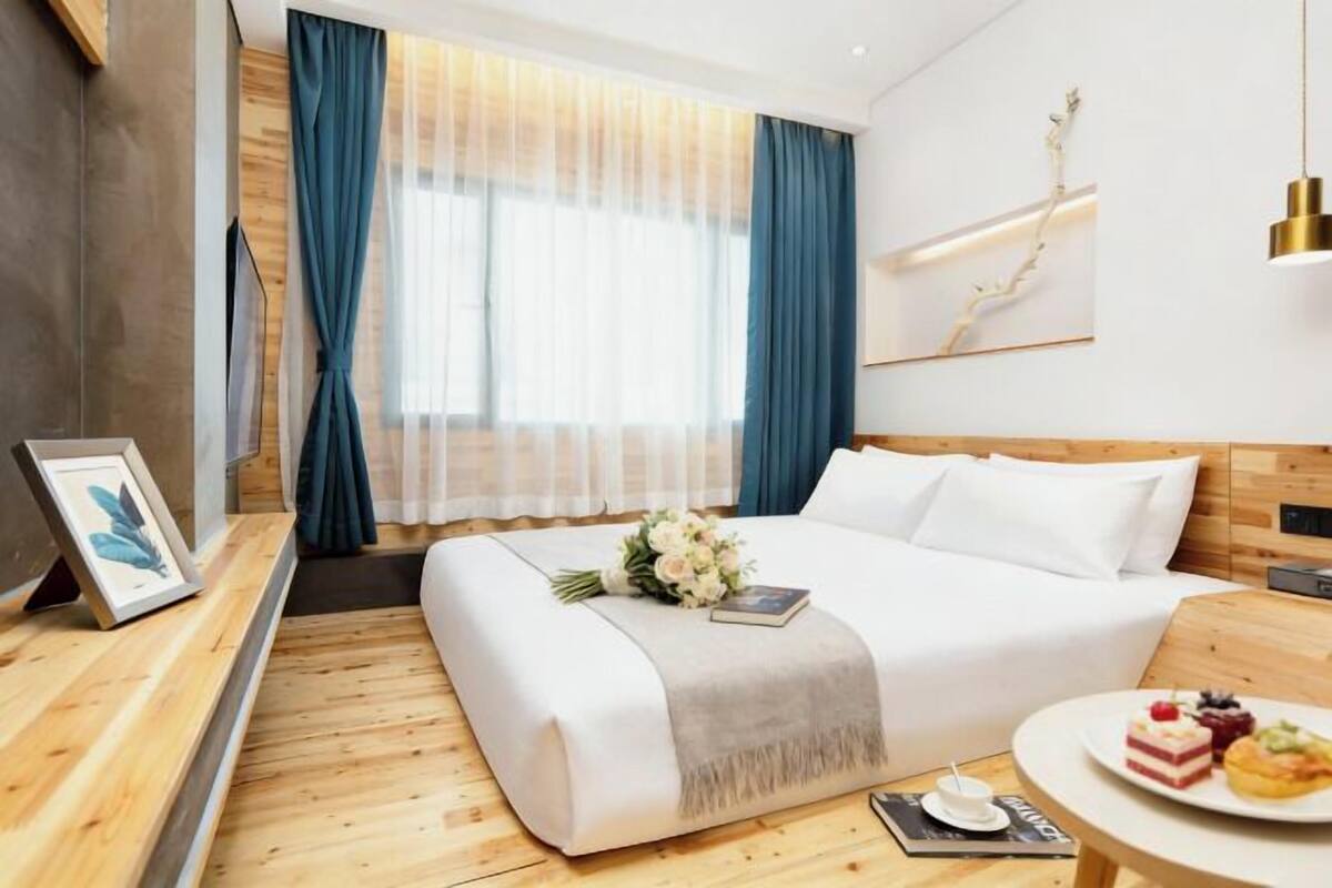 A modern hotel room with a large bed, a bedside table with a plate of pastries, a framed picture, and a window with blue curtains.