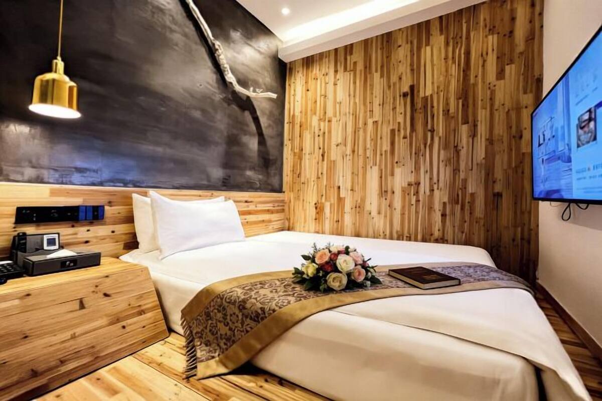 A modern hotel room with a wooden headboard, a flat-screen TV, a bedside table with a phone and a small device, and a neatly made bed with a floral arrangement.