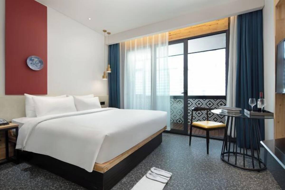 A hotel room with a bed, a chair, a small table with glasses, a TV, and a window with curtains.