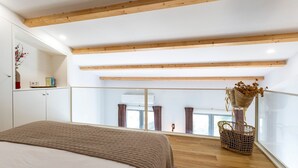 Un dormitorio con cama, vigas de madera, ventana con cortinas y balcón con cesta de mimbre.