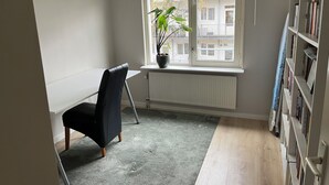 Een studeerkamer met een bureau, stoel, radiator, raam met plant en een boekenkast.