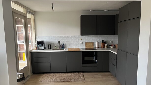 Een moderne keuken met donkergrijze kastjes, een wit aanrecht, een ingebouwde oven en een raam met uitzicht.