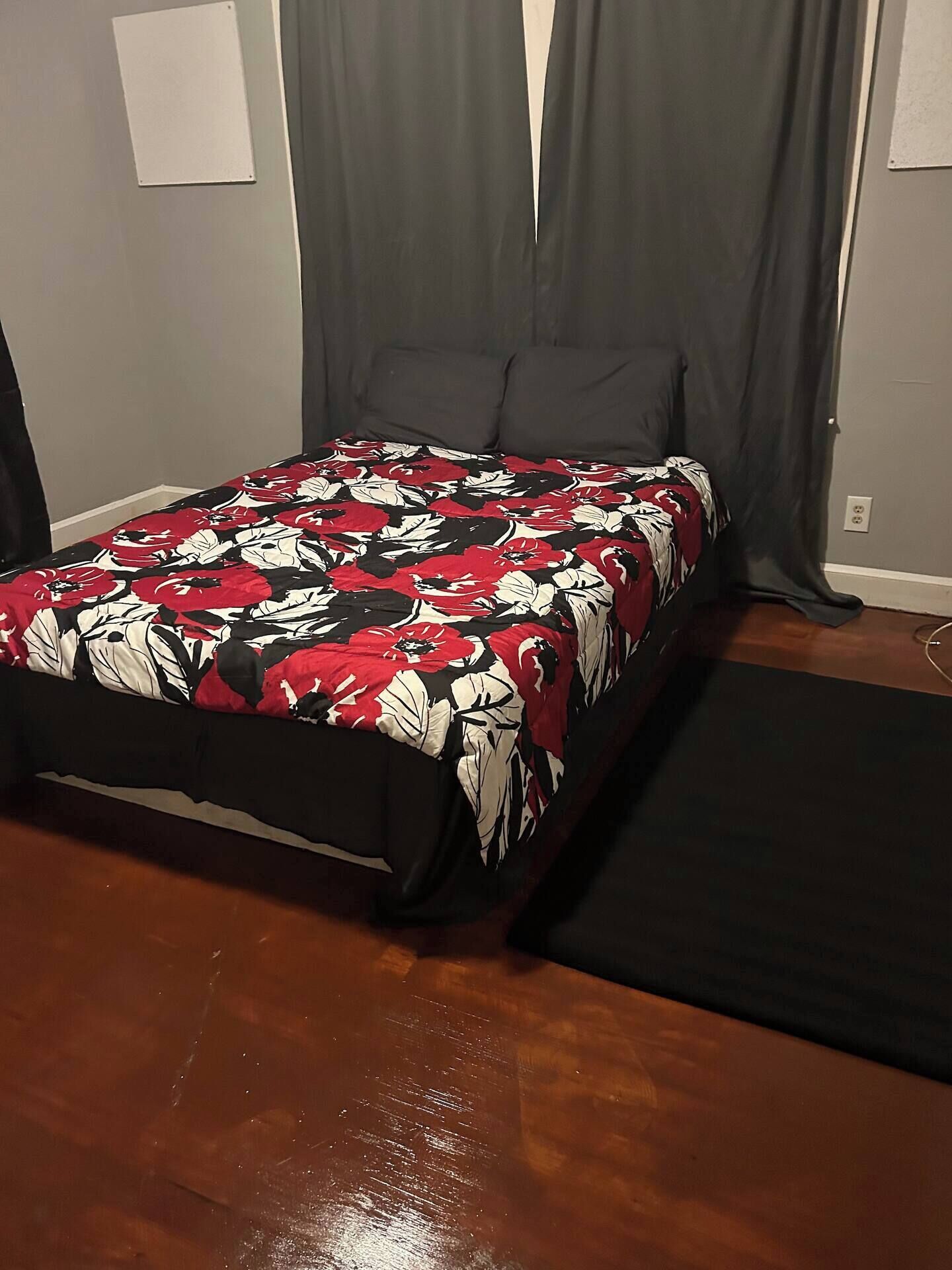 A bedroom with a bed, dark curtains, a wooden floor, and a small rug.