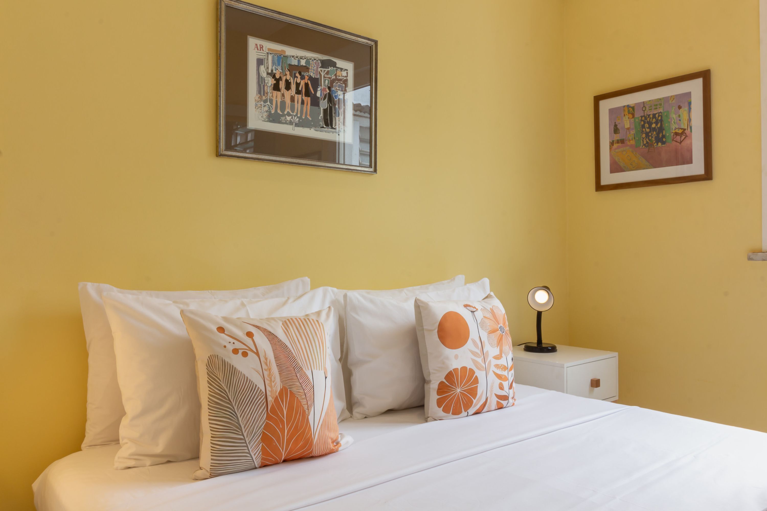 Een hotelkamer met een bed, kussens, een nachtkastje en twee ingelijste foto's aan de muur.