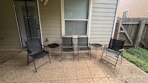 Een overdekt terras met vier zwarte metalen stoelen en een kleine ronde tafel, een schuifdeur en een raam met jaloezieën.