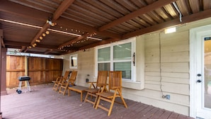 Un espacio exterior techado con piso de madera, cuatro sillas de madera, una parrilla y una ventana con persianas.