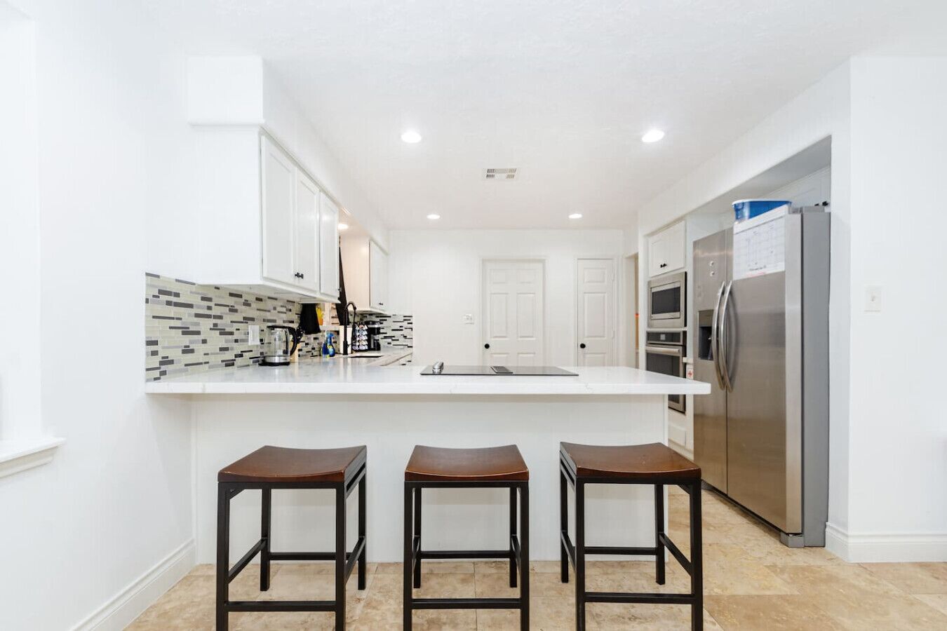 A modern kitchen with white cabinets, a tiled backsplash, a stainless steel refrigerator, and three wooden bar stools.