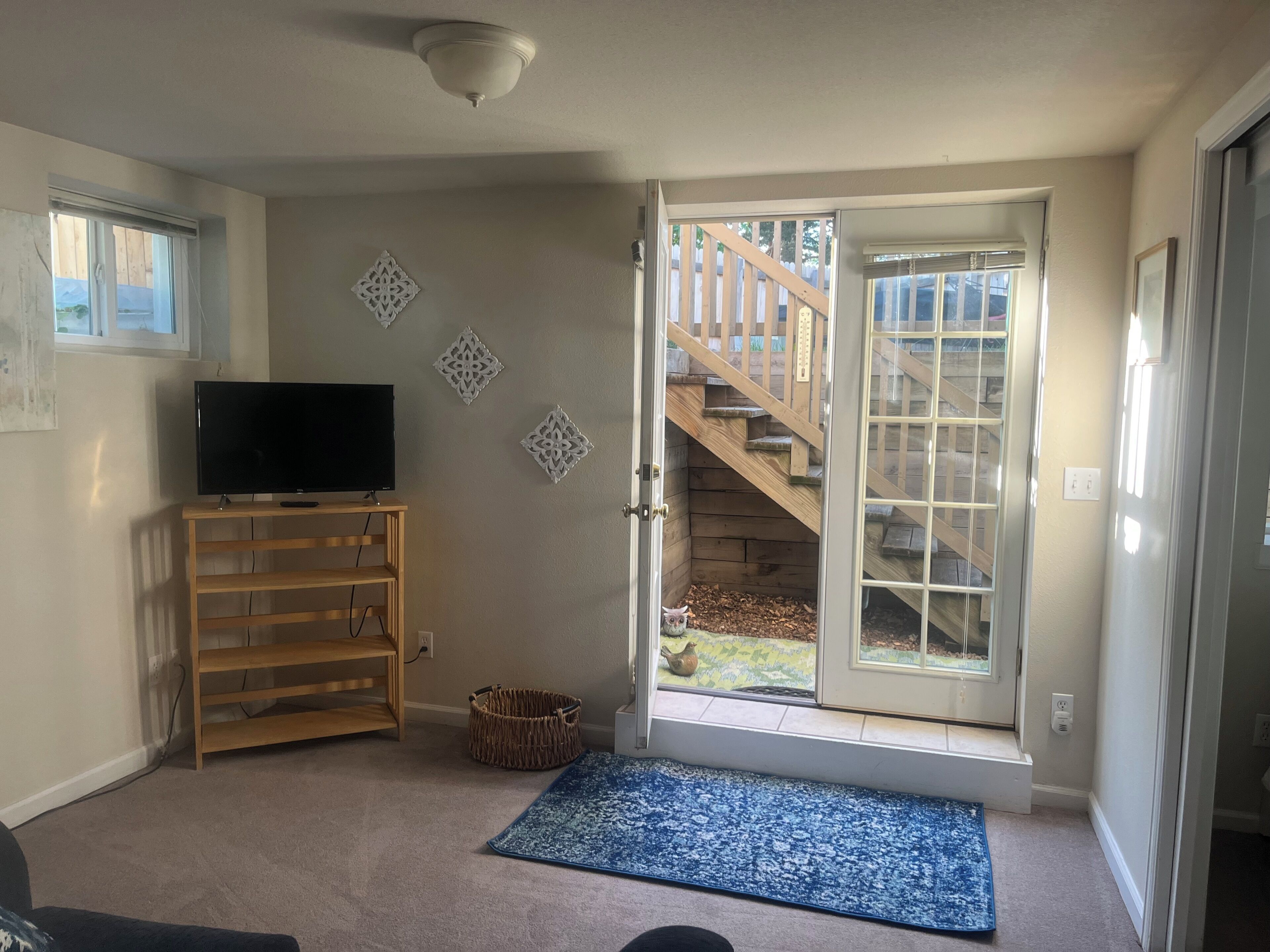 A living room with a TV on a wooden stand, a blue mat at the entrance, and a door leading to a wooden deck.
