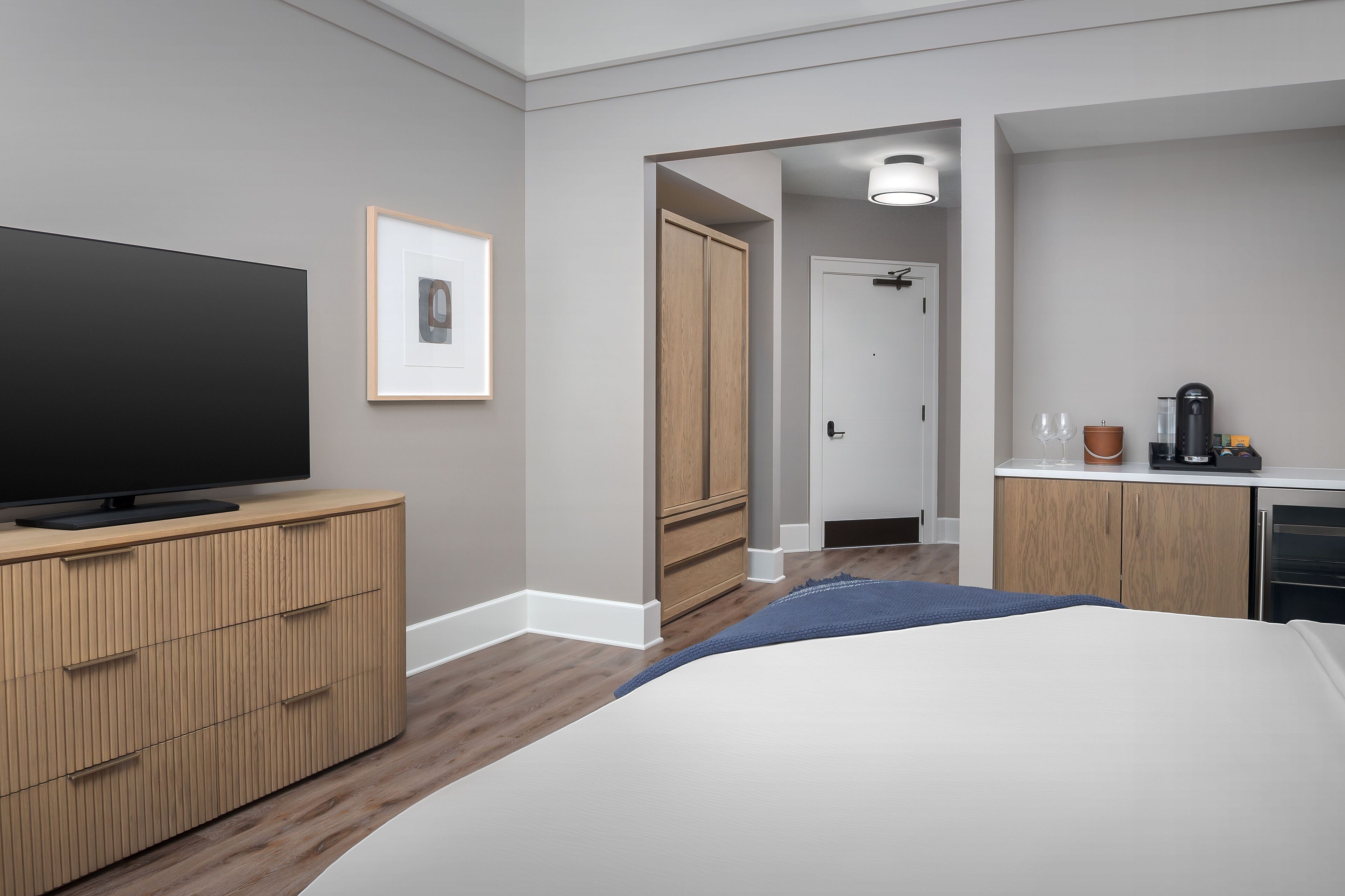 Quarto de hotel moderno com uma cama grande, televisão de tela plana, guarda-roupa de madeira e uma pequena cozinha.