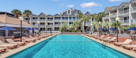 A swimming pool with lounge chairs and umbrellas, surrounded by multi-story buildings with balconies and palm trees.