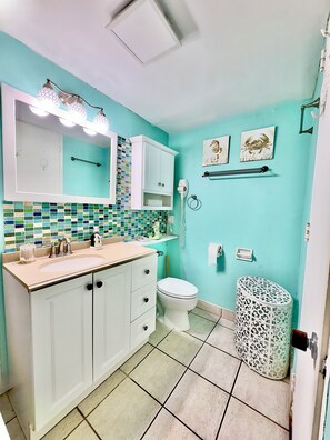 Ein Badezimmer mit türkisfarbenen Wänden, einem weißen Waschbecken mit Spiegel darüber, einer Toilette und einem Mülleimer mit Muster.