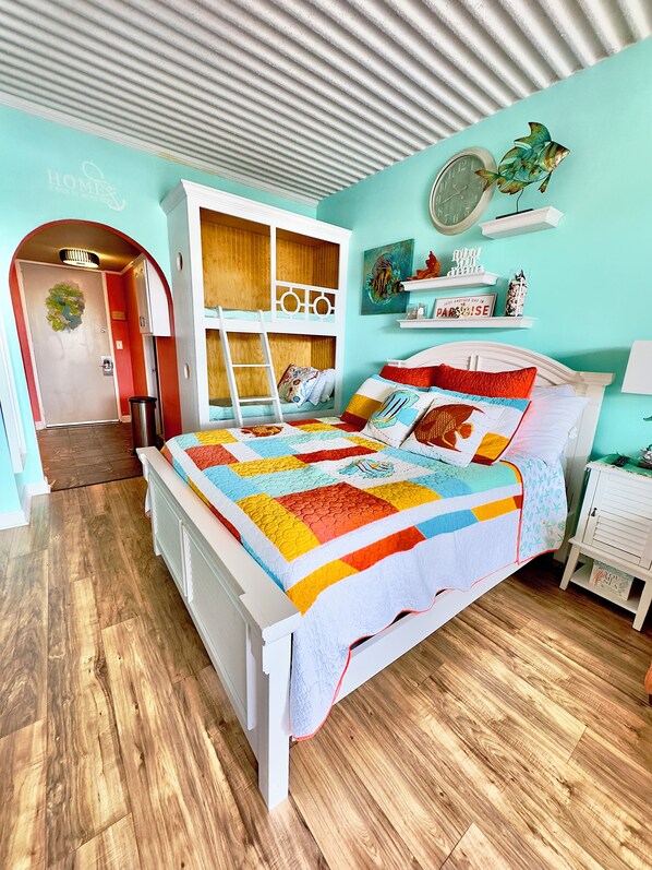 A bedroom with a wooden floor, a white bed with a colorful quilt, a bunk bed, a small desk, and a shelf with decorative items.