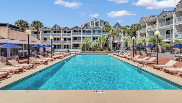 A swimming pool with lounge chairs and umbrellas, surrounded by multi-story buildings with balconies and palm trees. - NEW! 2nd Floor Studio w/Kitchen (Myrtle Beach)