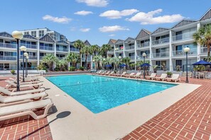 A swimming pool with lounge chairs and umbrellas, surrounded by a multi-story building with balconies and palm trees.