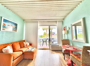 A room with a sofa, a small table, a wicker chair, a window, and a door leading to an outdoor area with a table and chairs.