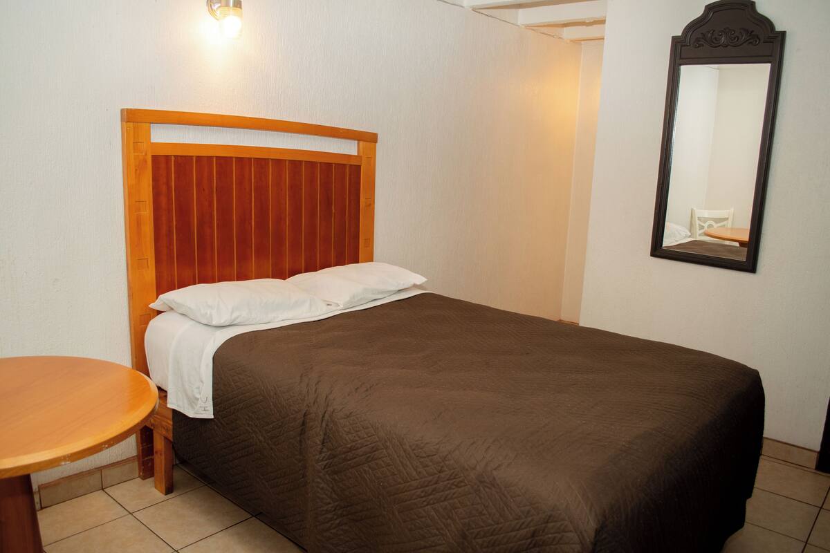 Uma cama de solteiro com cabeceira de madeira, uma mesa de madeira redonda e um espelho na parede.