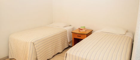 A small, clean room with two single beds, a nightstand with a book and a lamp, and a tiled floor.