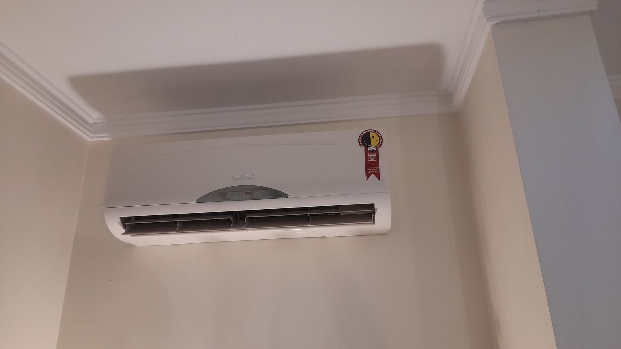 Un appareil de climatisation blanc, fixé au mur, avec un thermostat placé au plafond et un autocollant rouge portant une inscription.