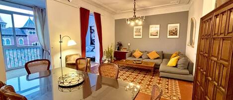 Una sala de estar con sofá, una mesa de centro y sillas. Se observa una araña, un ventanal con cortinas y una puerta con espejo.