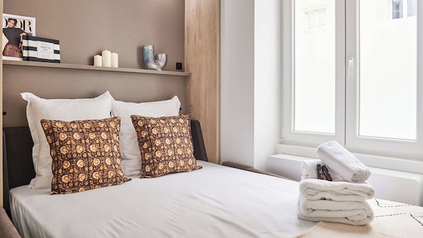 A neatly made bed with white linens and patterned pillows, a stack of white towels, and a magazine on a shelf. - Superb Studio Tour Eiffel/champ de Mars (Paris)