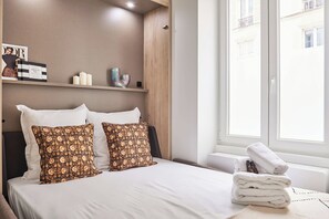 A neatly made bed with white linens and patterned pillows, a stack of white towels, and a magazine on a shelf. - Superb Studio Tour Eiffel/champ de Mars (Paris)