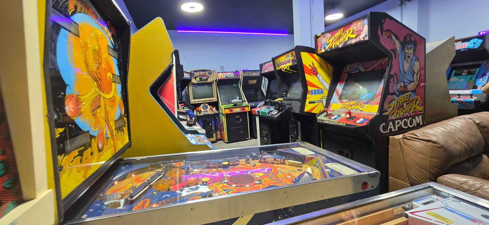 A row of arcade machines with colorful graphics and a pinball table in the foreground.