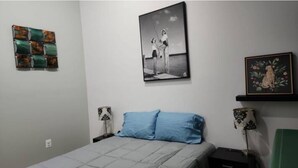 A bedroom with a bed, a wall-mounted picture, a shelf with a framed picture, and a decorative wall piece.