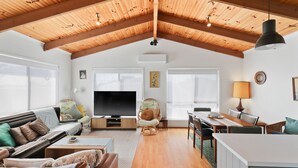 Un amplio espacio de sala con techo de madera alto, un televisor de pantalla plana, una mesa de comedor con sillas y una zona de estar acogedora con cojines.