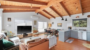 Amplia sala de estar con televisor grande, vigas de madera en el techo, comedor y cocina con vista al exterior.