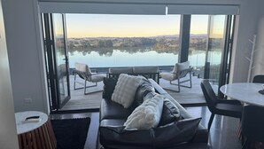 A modern living room with a large glass door leading to a balcony overlooking a lake. - Estuary Views - Riverhead Holiday House (Auckland)