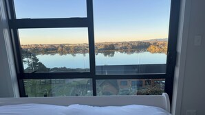 A bedroom with a large window overlooking a lake and surrounding landscape. - Estuary Views - Riverhead Holiday House (Auckland)