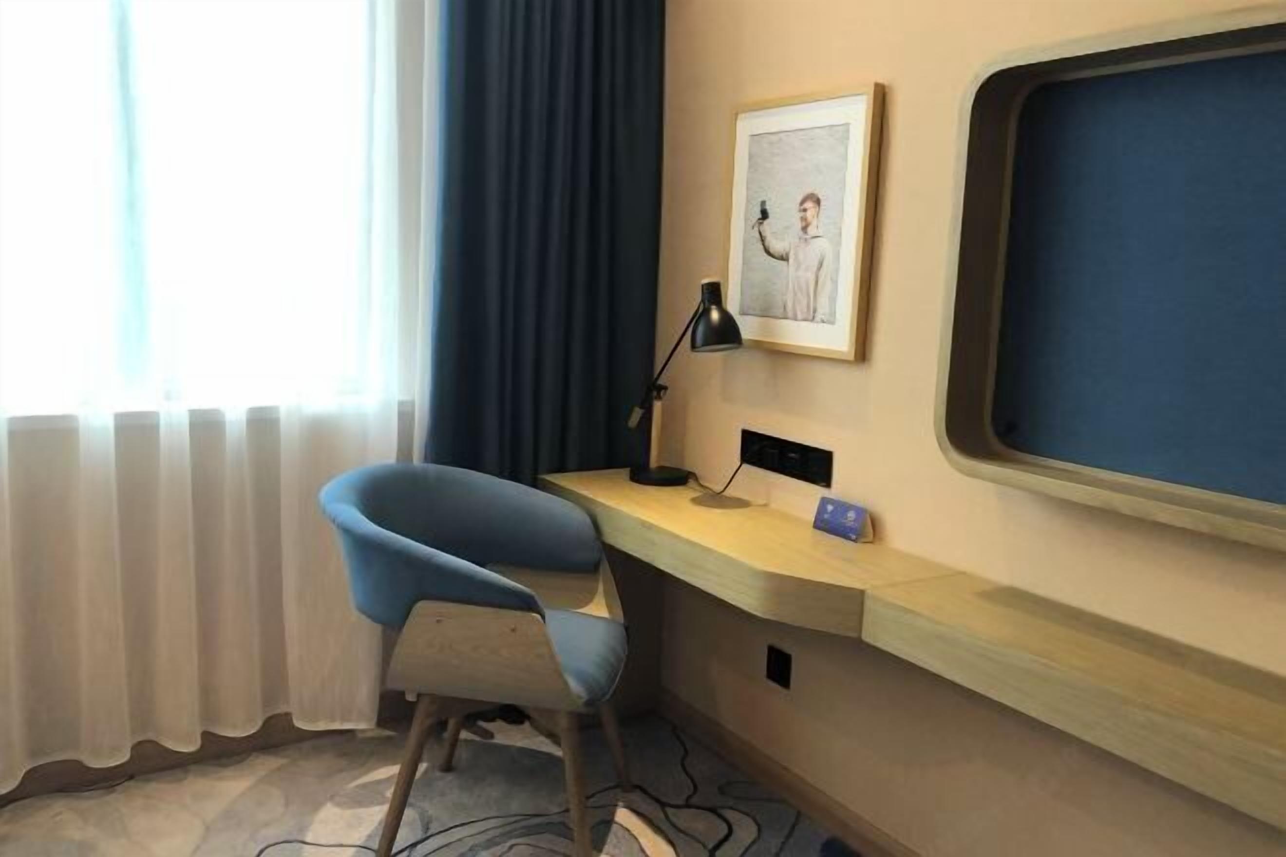 A hotel room with a desk, chair, window with curtains, a framed picture, and a large window.