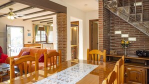 Un comedor con una mesa de madera, sillas y un mantel de encaje. Se observa una escalera con un muro de ladrillo y una puerta que da a otra habitación.