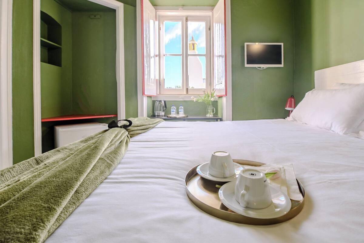 Une chambre d’hôtel avec un lit, une fenêtre avec des rideaux, un plateau avec des tasses et des soucoupes, et un téléviseur fixé au mur.